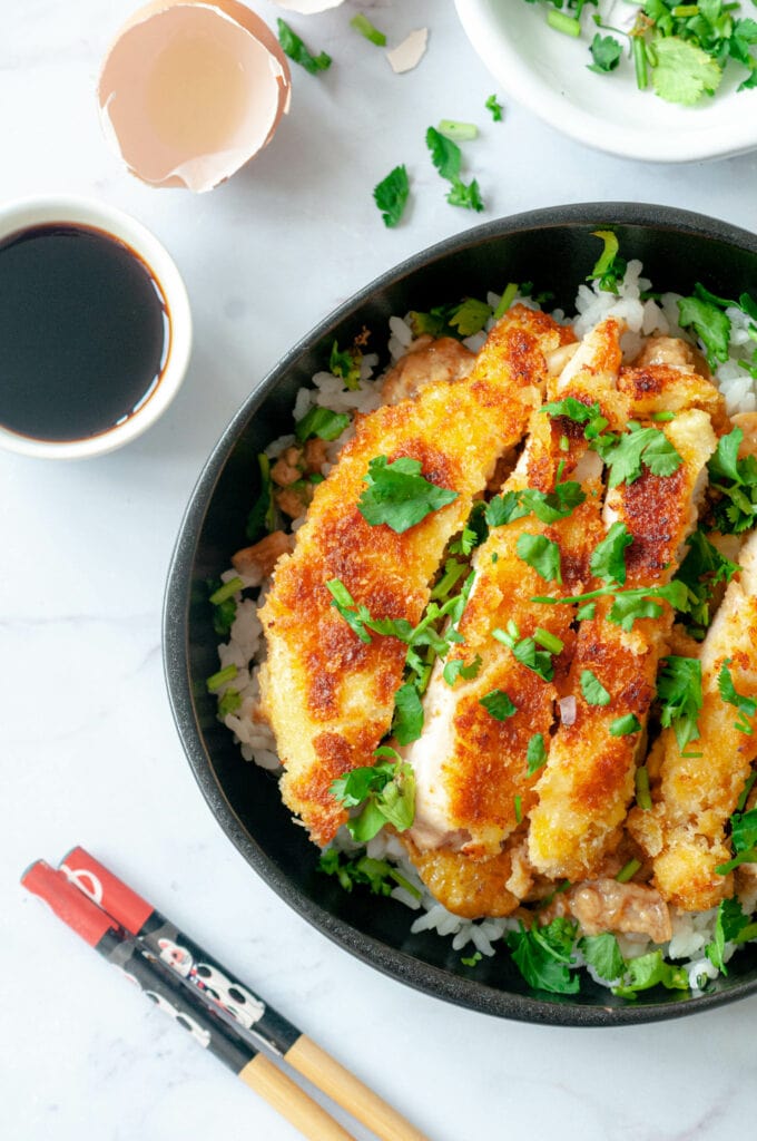 Zoom sur le Katsudon. À côté : des baguettes, un bol de sauce soja, un de coriandre et une coquille d'oeuf.