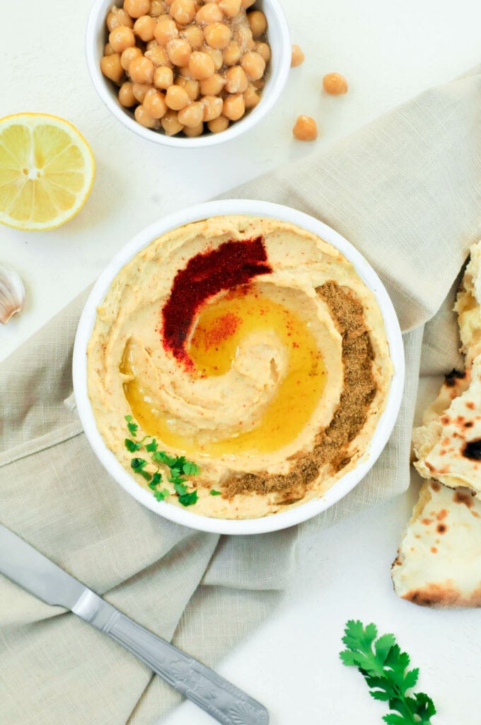 Houmous dans un bol avec du cumin, du paprika et de l'huile d'olive. Autour, des pitas, du citron et un petit bol de pois chiches.