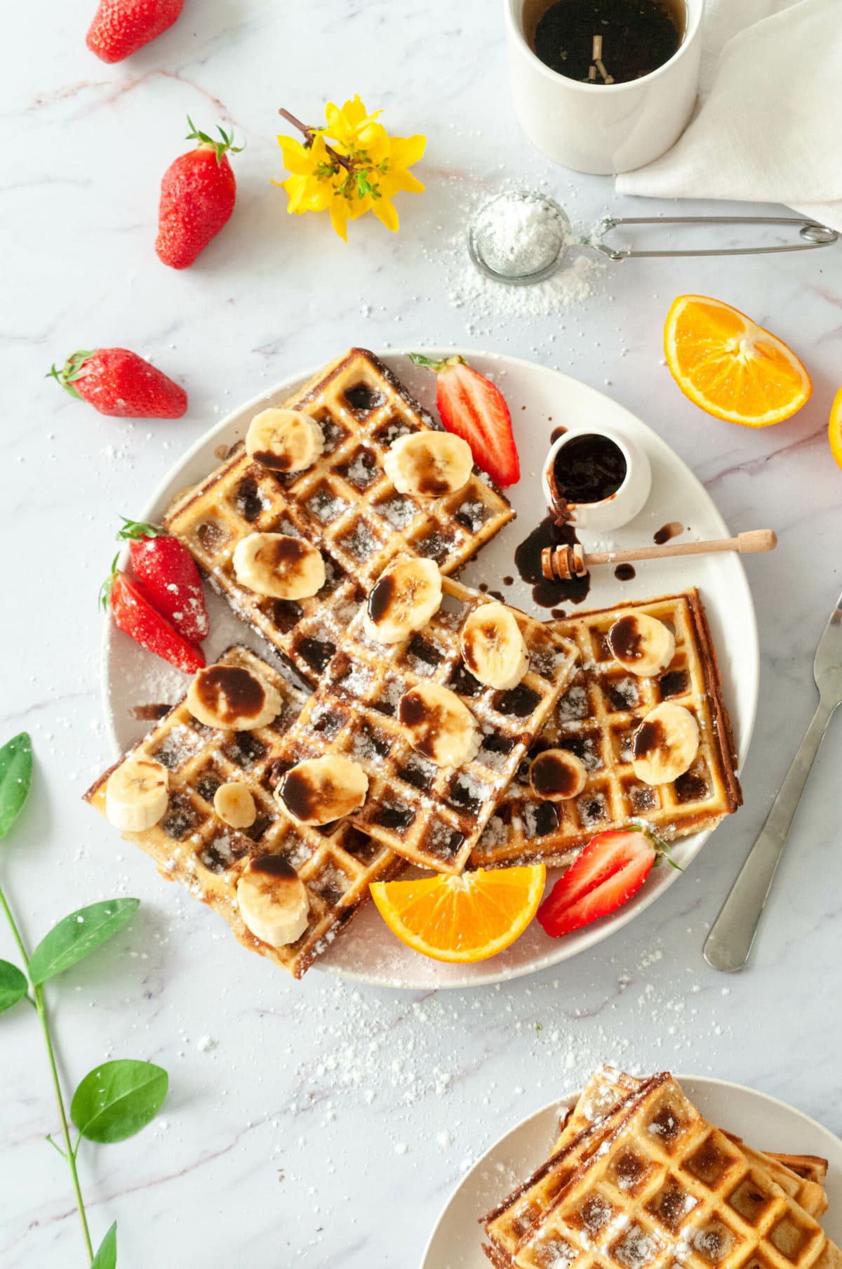 Several waffles on a plate with melted chocolate, hot chocolate, strawberries and orange wedges.