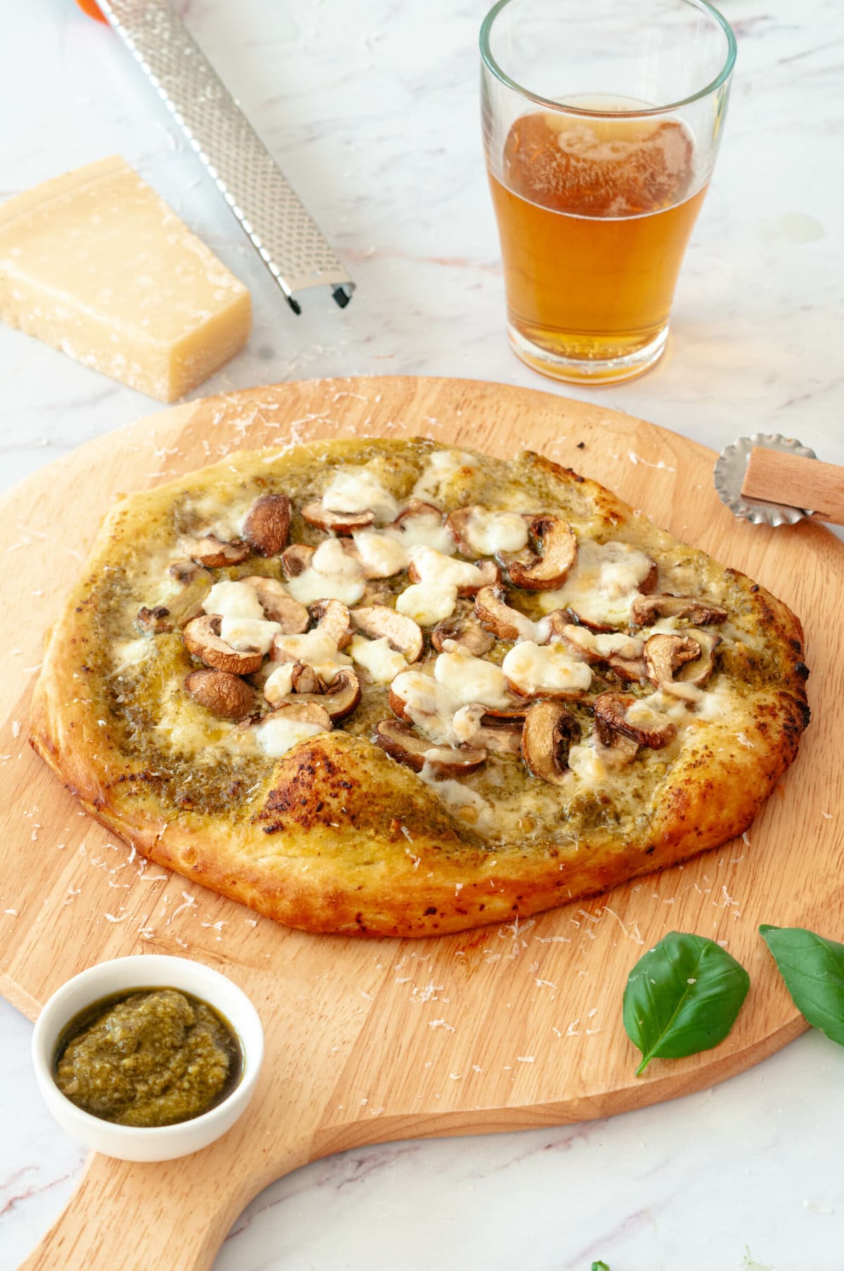 Pizza sur une planche avec un morceau de Parmesan, un bol de Pesto et un verre de bière.