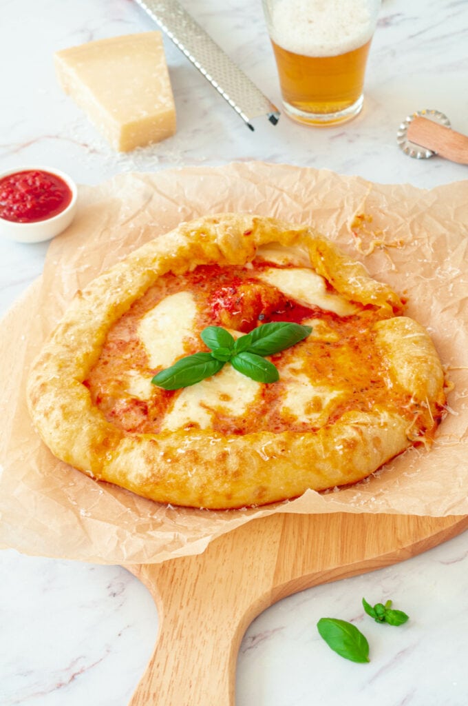 Pizza Margherita sur une planche avec un morceau de Parmesan, un bol de sauce tomate et un verre de bière.