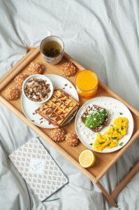 Plateau de brunch dans un lit avec avocado toast, gaufres, jus d'orange, thé et chouquettes.