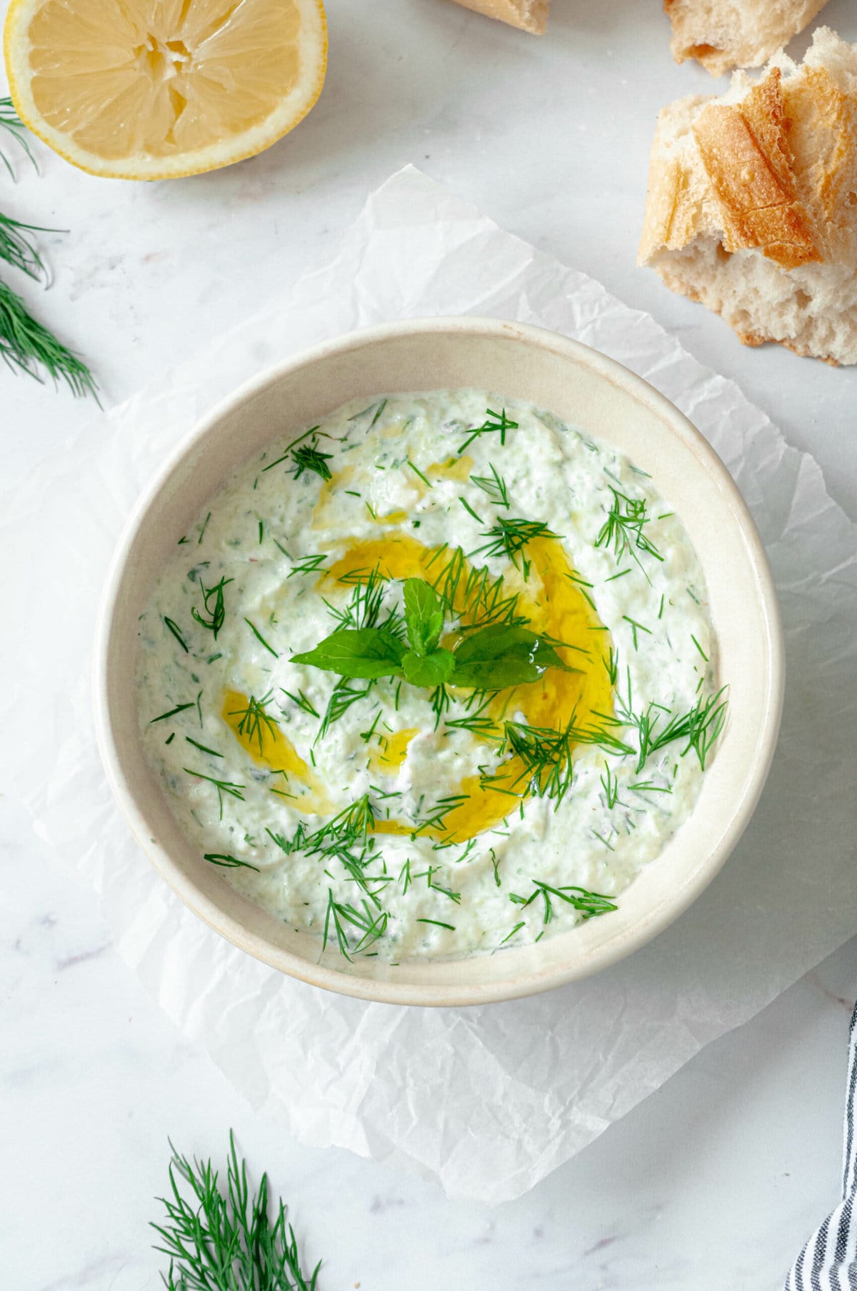 Tzatziki in a beige bowl on parchment paper with a drizzle of olive oil, fresh dill and mint as a topping.