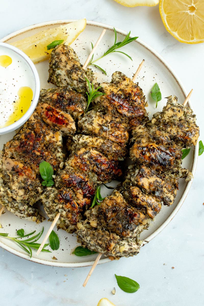Chicken skewers on a plate with herbs and lemon wedges.