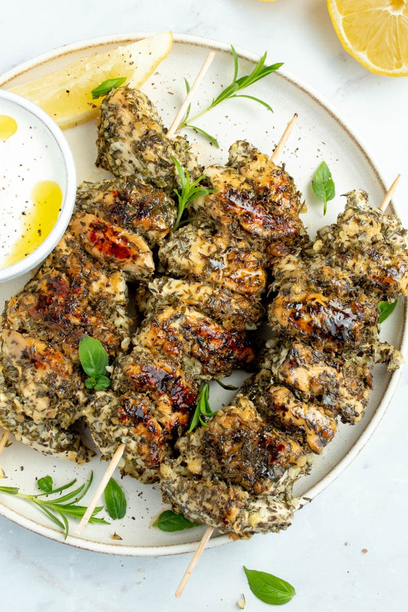 Chicken skewers on a plate with herbs and lemon wedges.