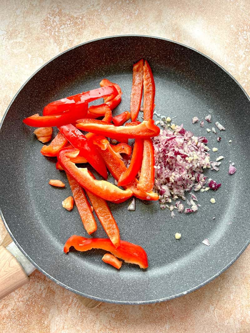 Sliced red peppers in a frying pan with garlic and red onion.