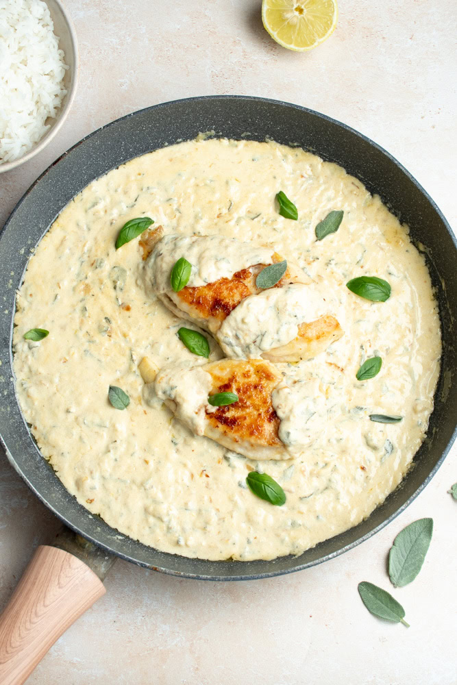 Two golden chicken fillets bathed in a pan of lemon-sage cream sauce, with fresh basil leaves.