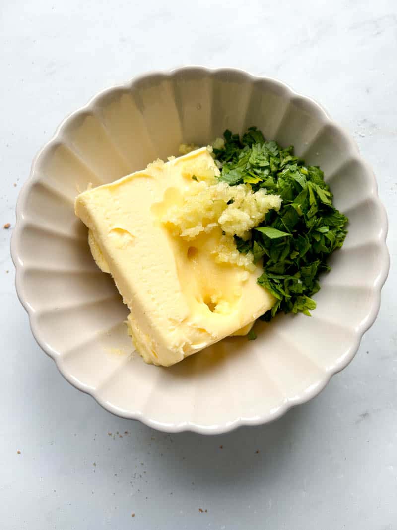 Soft butter, pressed garlic and chopped fresh parsley in a beige bowl.