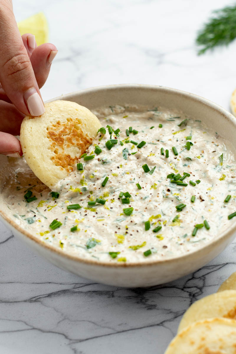 Tuna dip in a bowl with a hand dipping a blini in it.