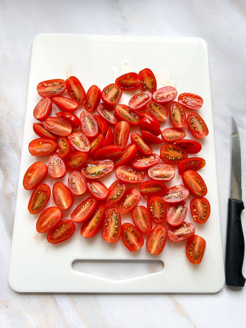 Cherry tomatoes cut in half on a white cupping board, with a knife.