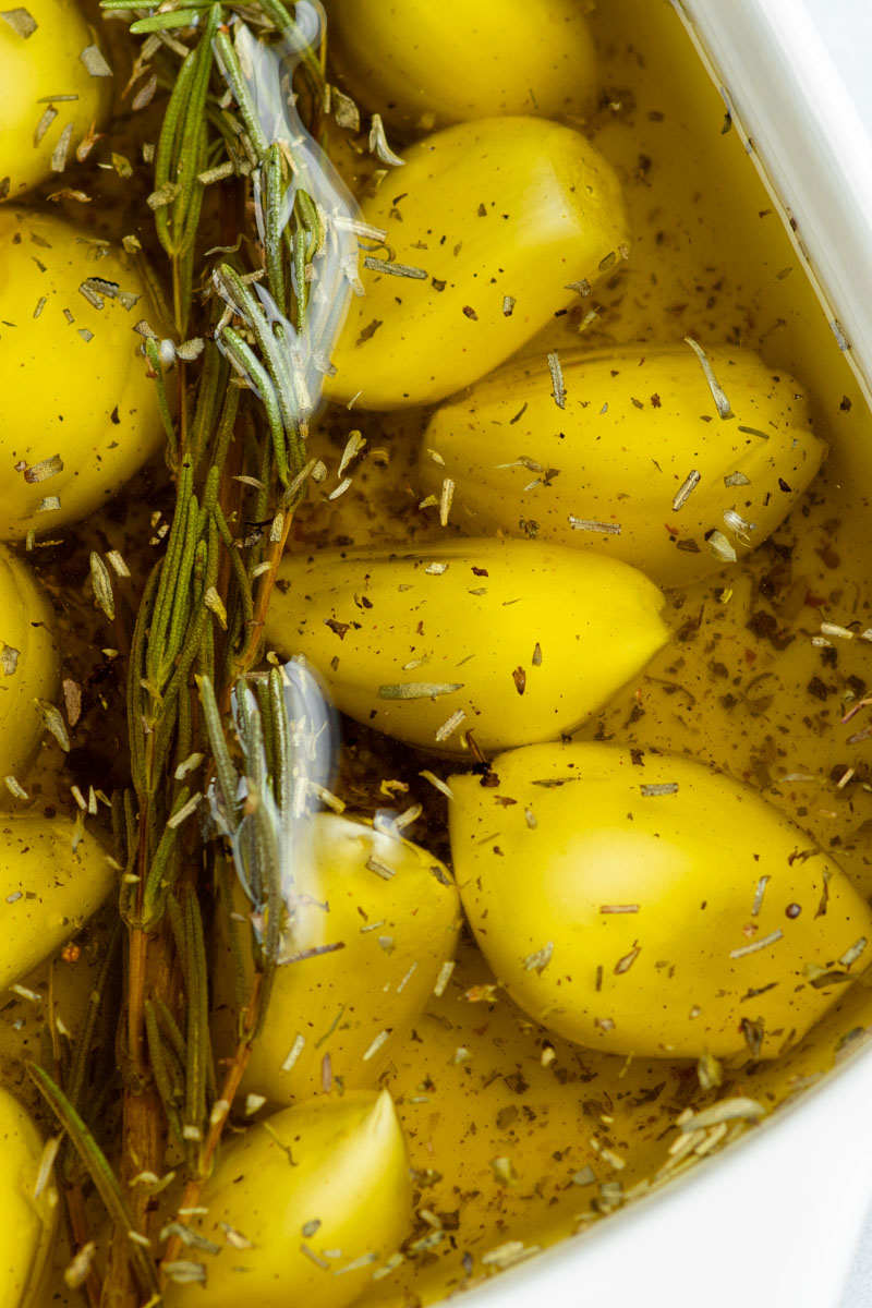 Fresh garlic cloves in an oven-safe dish submerged with olive oil, with a rosemary sprig.