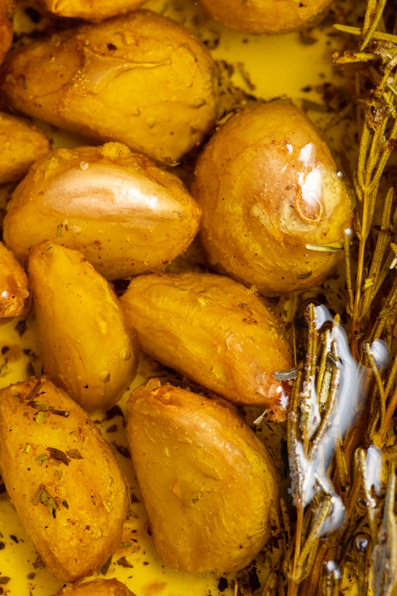 Confit garlic cloves in a white oven-safe dish with olive oil and one rosemary sprig.