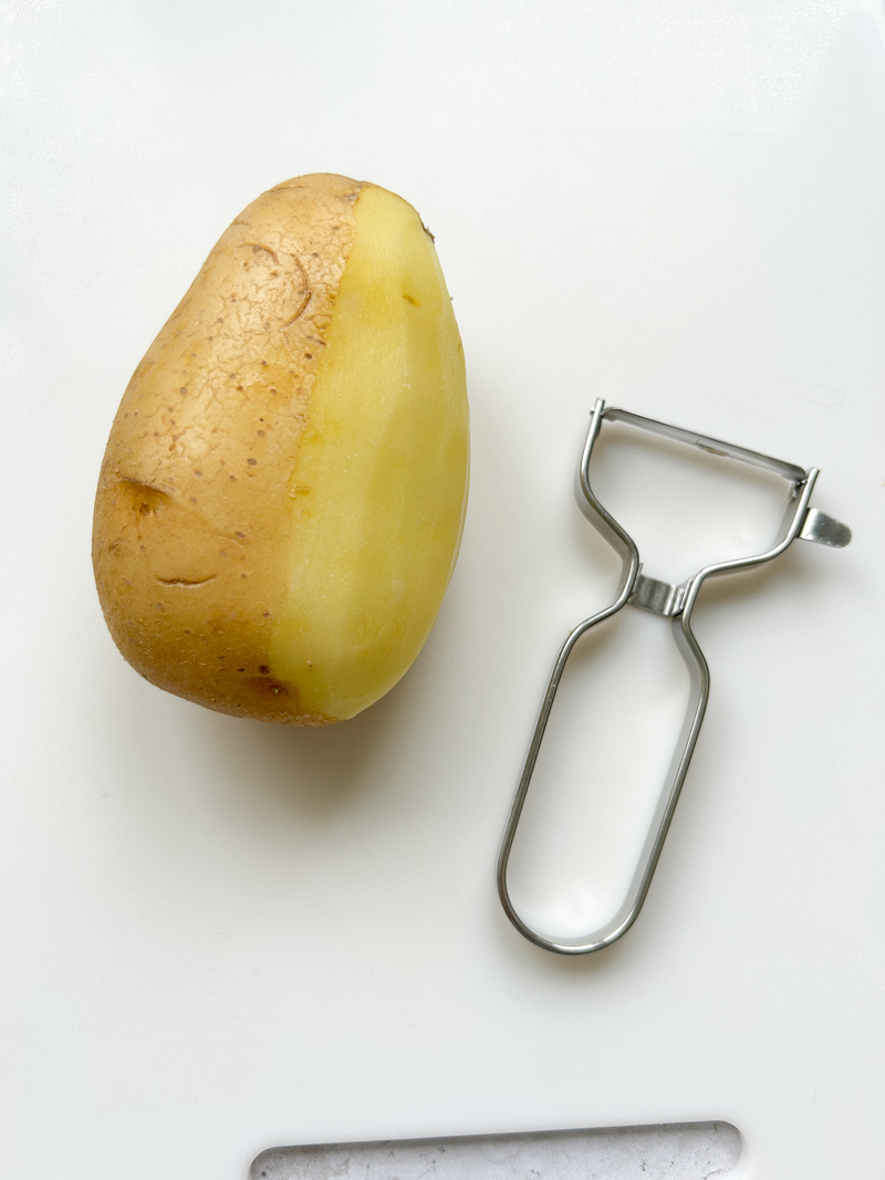 Pomme de terre à moitié épluchée sur une planche à découper blanche, avec un éplucheur à côté.