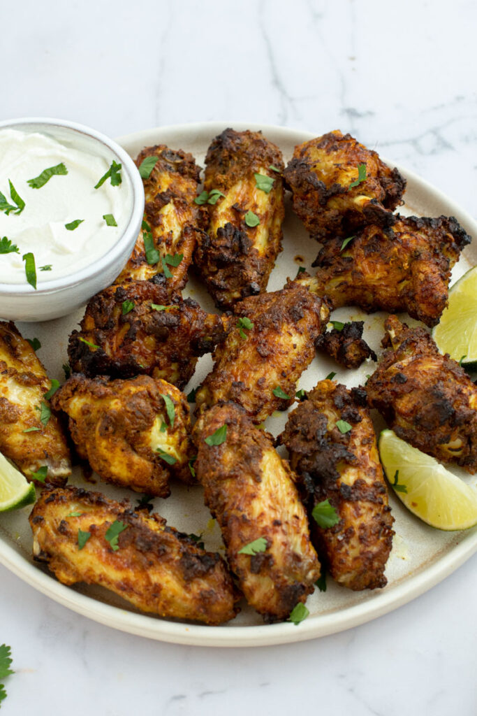 Tandoori chicken wings in a white plate with lime edges and a bowl of white sauce.