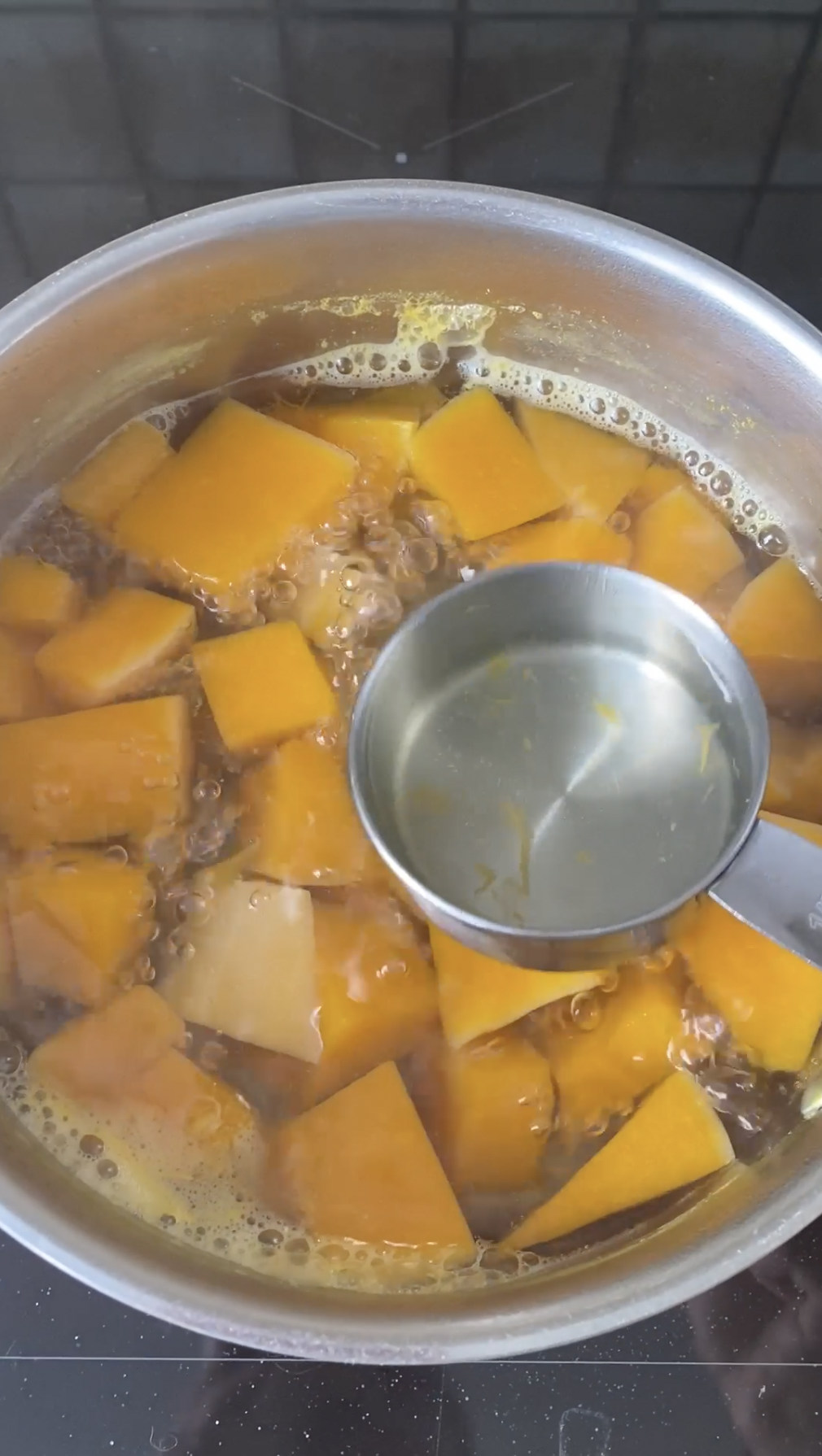 A cup hold by a hand reserving some cooking water in a pot with butternut squash cubes.