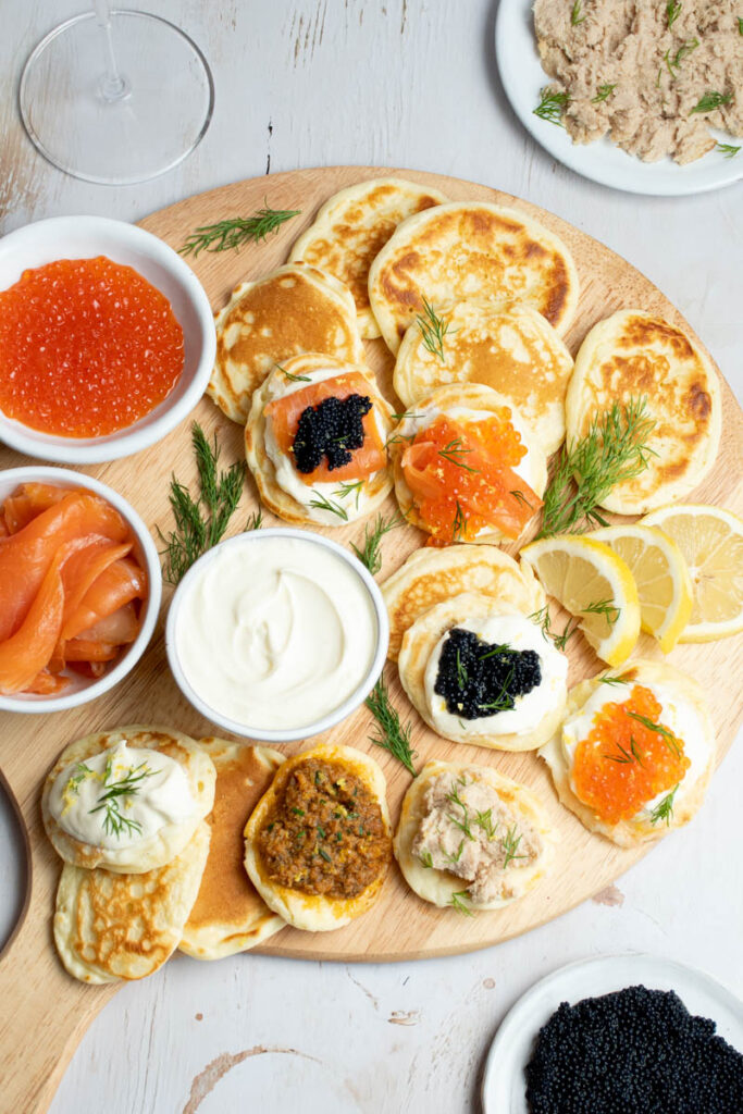 Lots of blini on a round wooden board, with many different toppings.