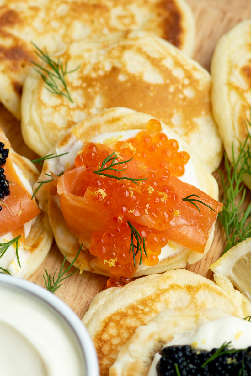 Lots of blini on a round wooden board, with many different toppings. Focus on one of the blini, with creme fraiche, smoked salmon and salmon roe.