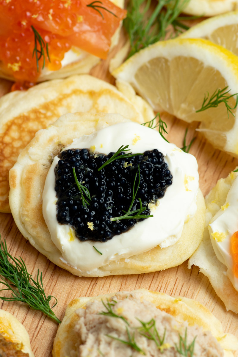 Lots of blini on a round wooden board, with many different toppings. Focus on one of the blini, with creme fraiche and caviar.