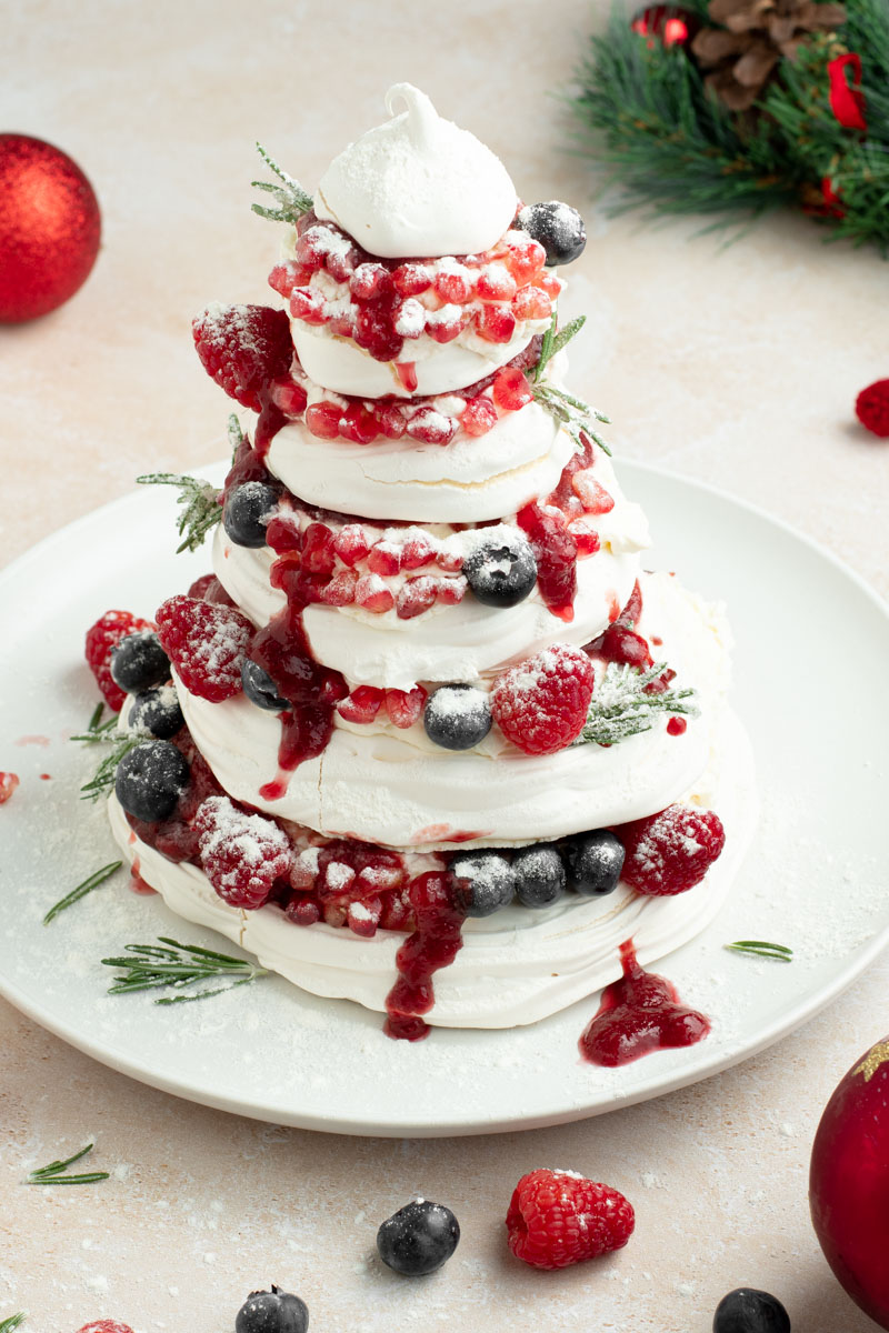 Pavlova de Noël dans une assiette blanche avec des fruits rouges, une boule de Noël et une couronne de sapin.