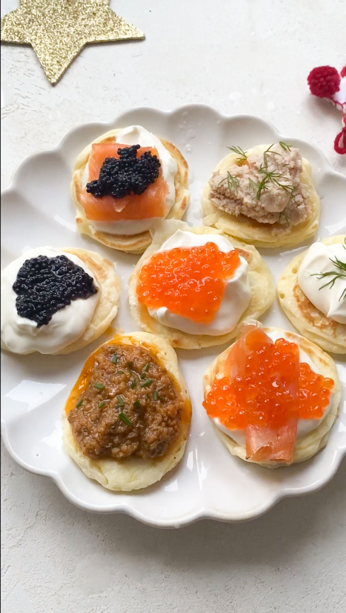 Seven blini placed on a white plate, garnished with different toppings.