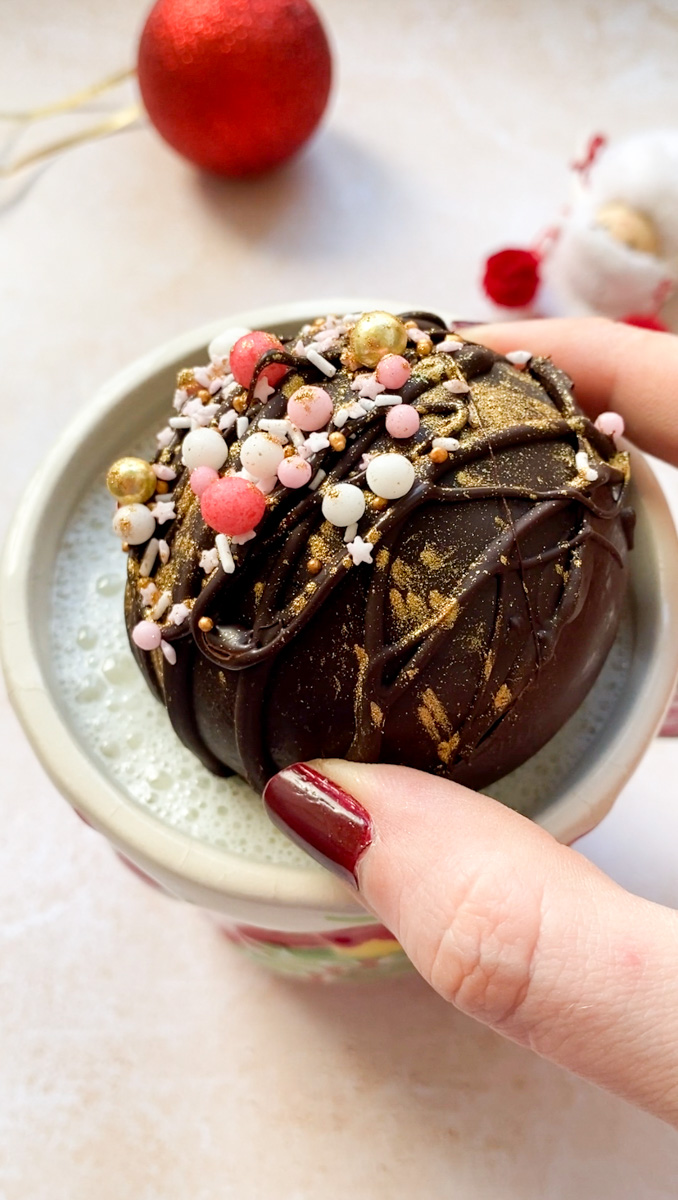 Chocolate bomb hold by a hand and placed in a cup of milk.