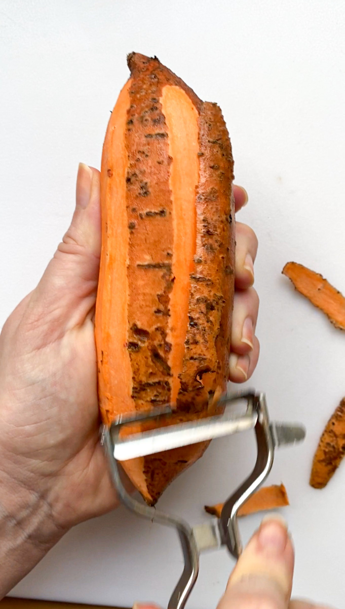 One hand holding a sweet potato and the other one holding a vegetable peeler, peeling the sweet potato.