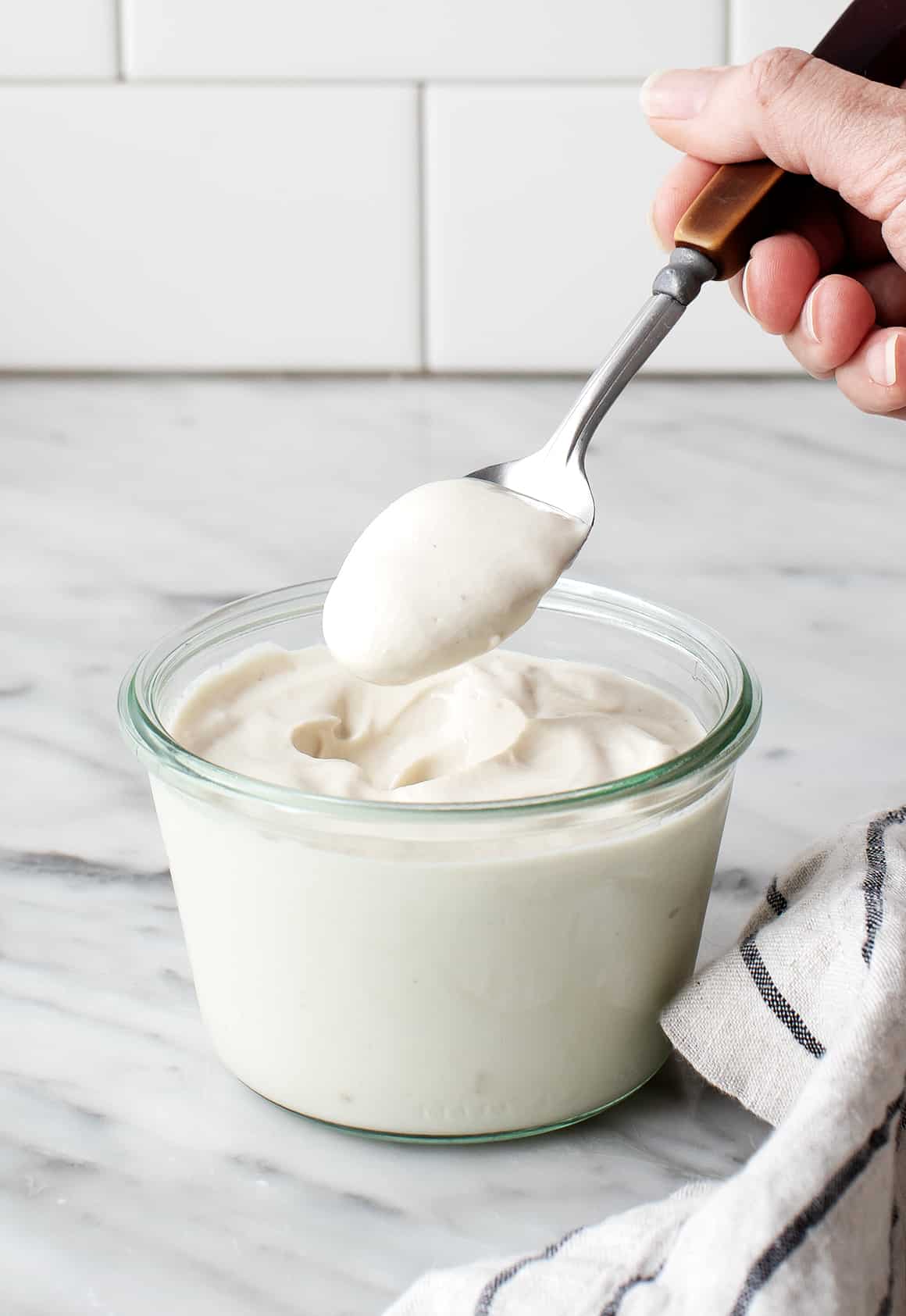 A hand holding a spoon that pour vegan sour cream back into the jar of sour cream.