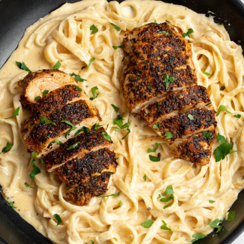 Blackened chicken on top of creamy Alfredo pasta in a black frying pan, with chopped fresh parsley.