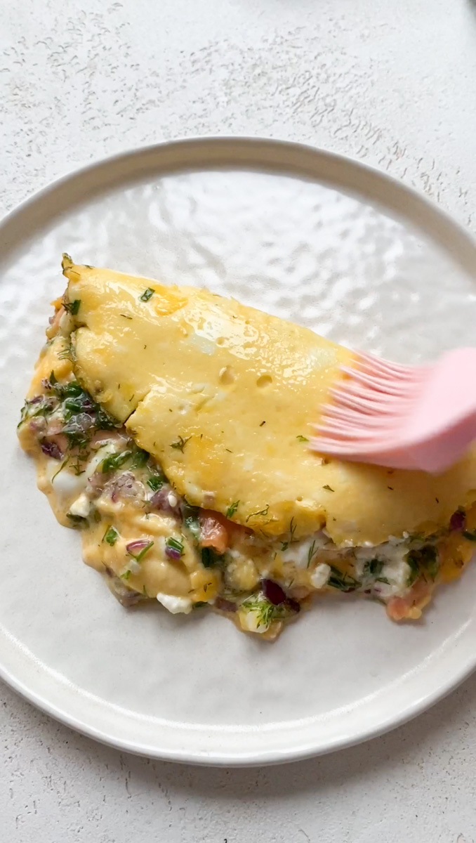 A pink silicone brush brushing melted butter on the top of the cooked omelette, placed on a white plate.