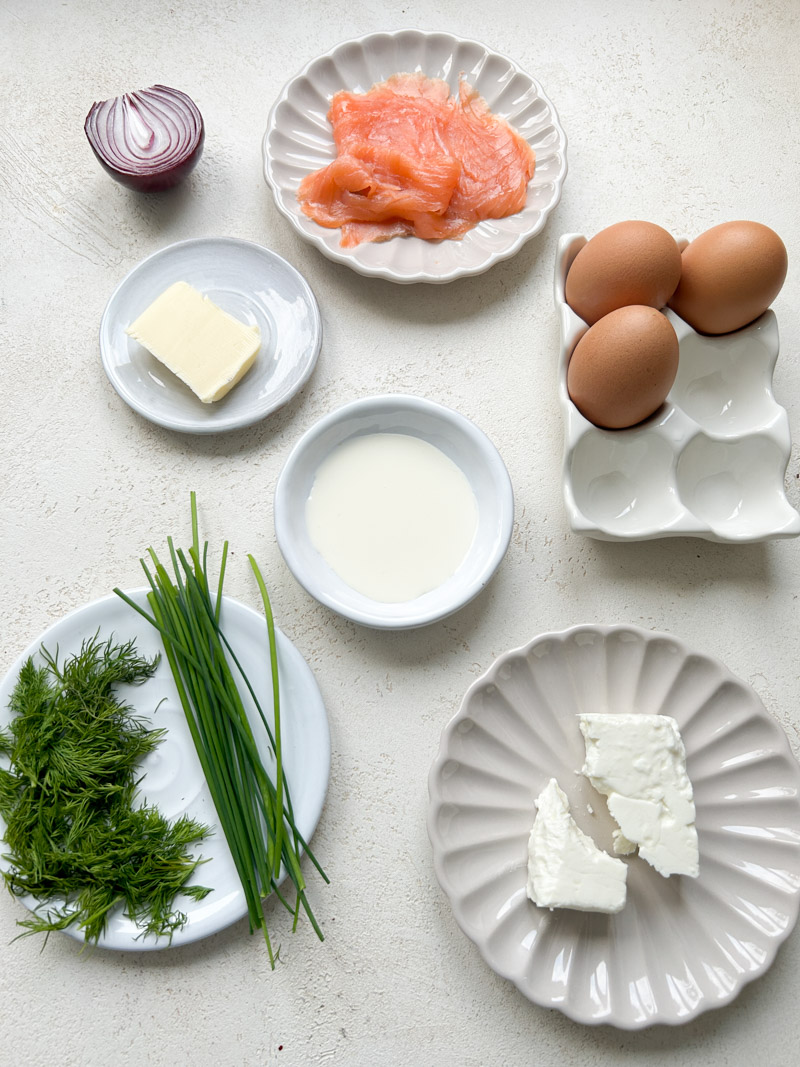 Les ingrédients pour faire une omelette avec du saumon, des herbes, de la feta et de l'oignon rouge dans des bols blancs et beiges.
