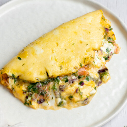 Smoked salmon omelette in a white plate, with feta cheese, red onion and fresh herbs.