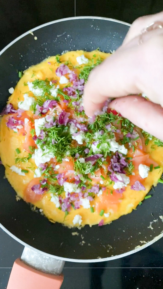 Morceaux de saumon fumé, feta émiettée, oignon rouge coupé en dés et herbes fraîches hachées ajoutés par une main dans la poêle de l'omelette.