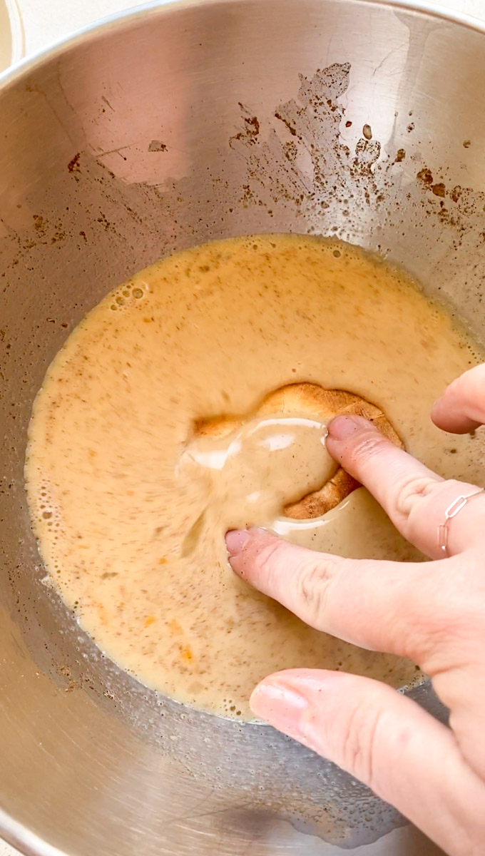 A hand dipping half a bagel into the batter.