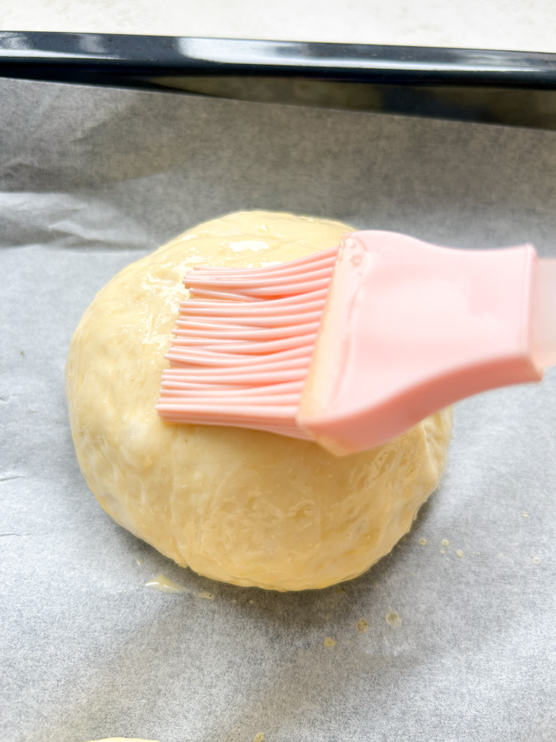 A pink silicone brush is brushing one ball of dough with the milk and egg mixture.