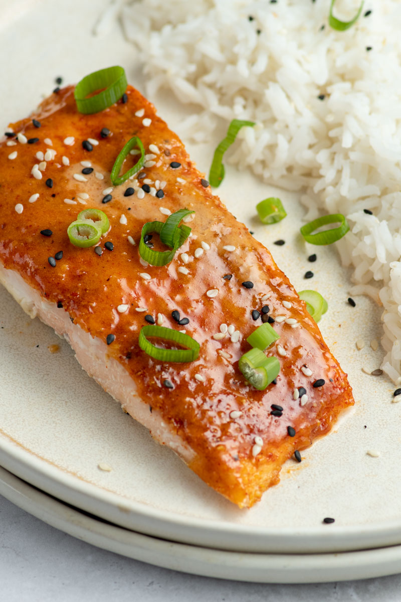Bang Bang salmon in a beige plate with white rice, sliced green onions and sesame seeds.