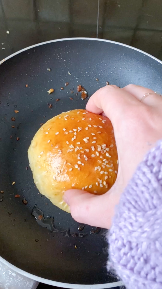 Une main tient un pain à burger, qui cuit dans une poêle.