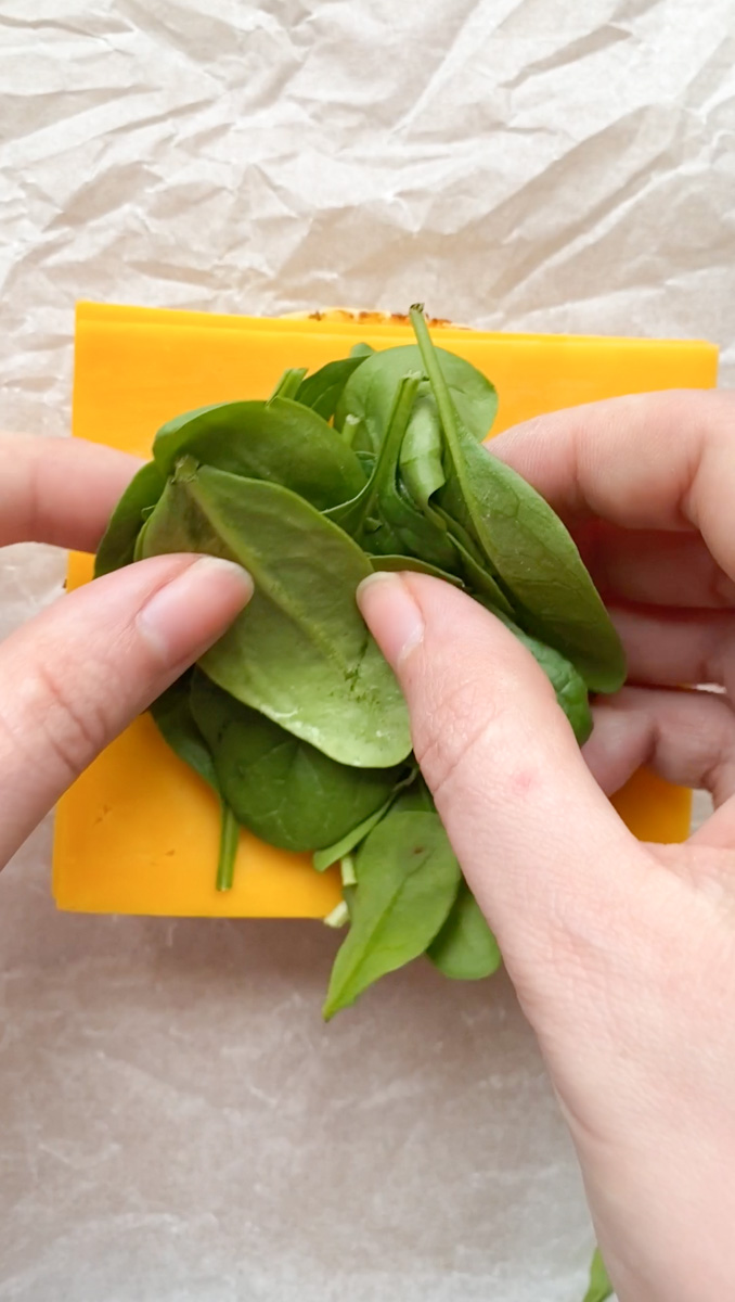 Two hands placing baby spinach on top of the cheddar cheese slices.