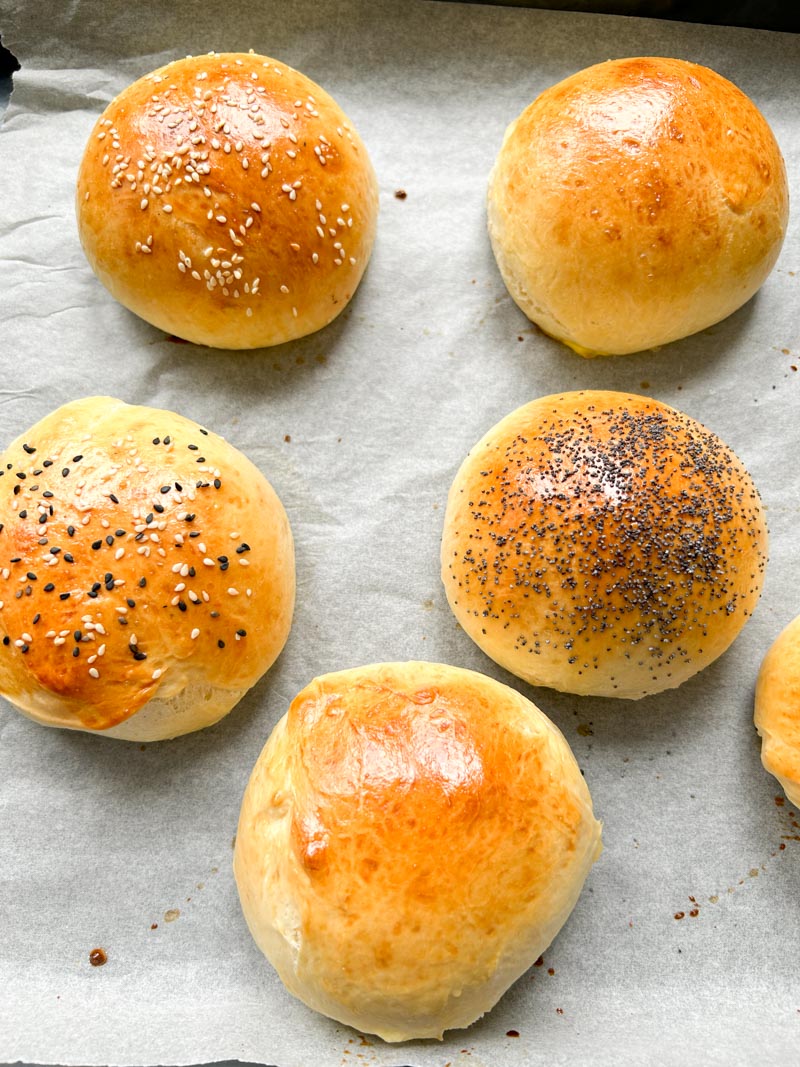 Baked brioche burger buns on a baking tray lined with parchment paper, with different toppings - sesame seeds, poppy seeds, etc...