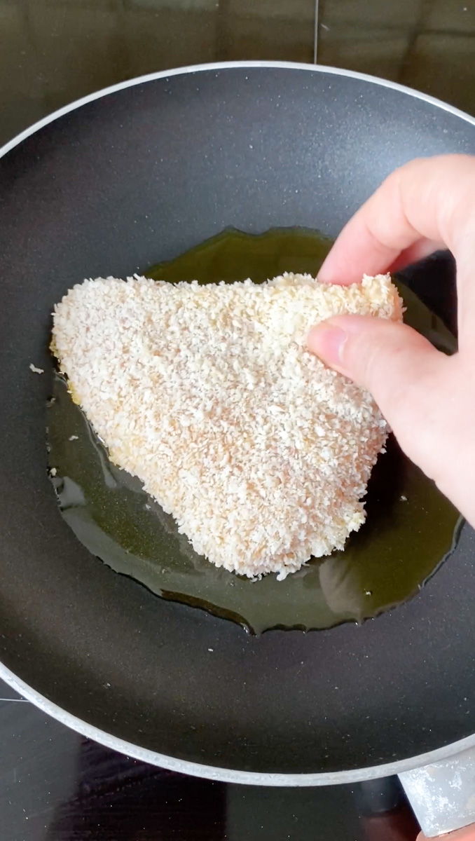 A hand placing coated chicken breast in a pan with olive oil.
