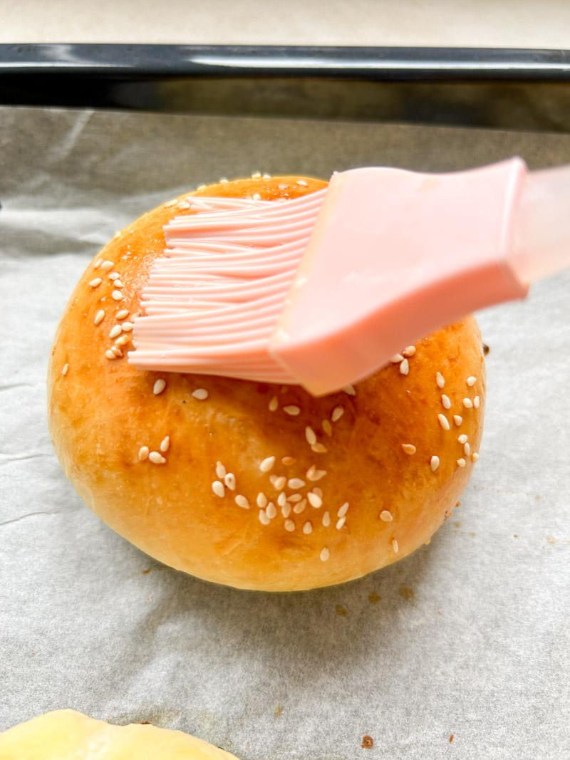 Un pinceau en silicone rose badigeonne un pain à burger cuit avec le mélange de lait et d'œuf.