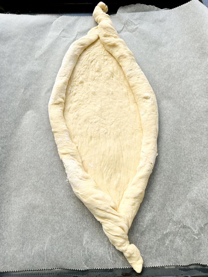 The Khachapuri's dough, in a boat shape, is placed  on an oven tray lined with parchment paper.