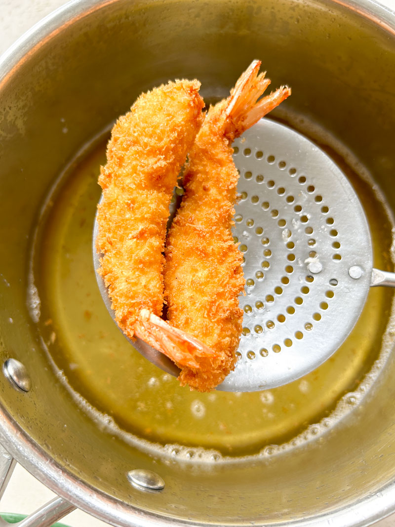 Two Ebi Fry cooked and held by a skimmer over a pan of boiling oil.