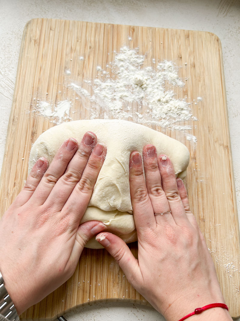 Two hands kneading the dough on a lightly floured wooden cutting board.