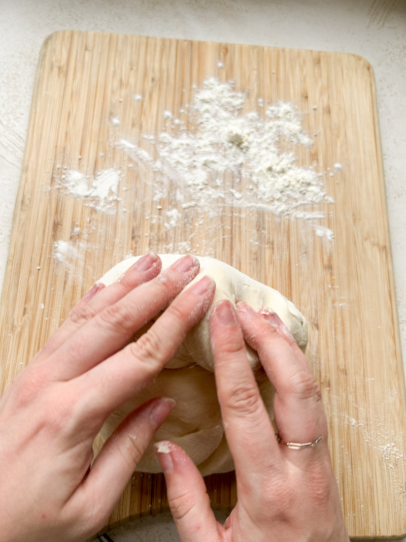 Two hands kneading the dough on a lightly floured wooden cutting board.
