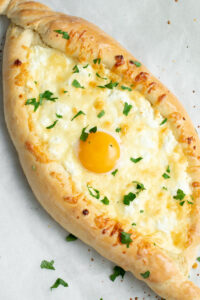 Khachapuri - pillowy and fluffy bread filled with melted cheeses and a runny egg yolk - on parchment paper with fresh chopped parsley.