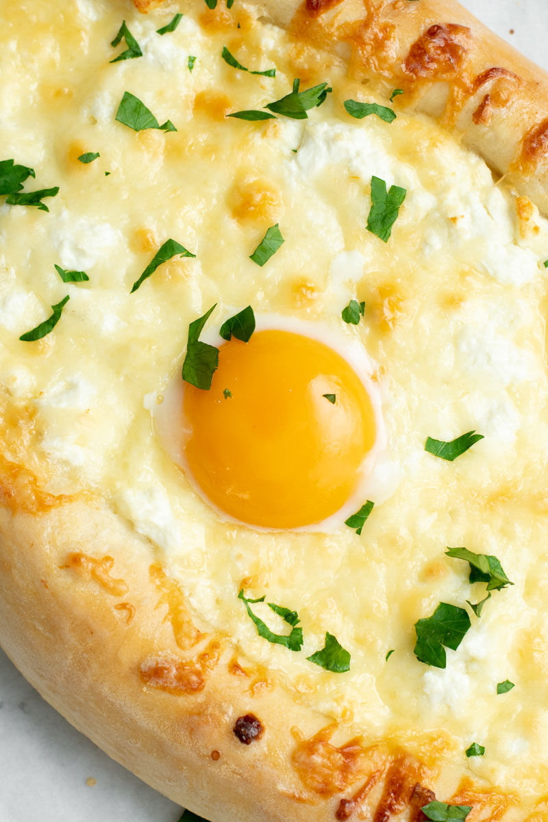 Khachapuri - pillowy and fluffy bread filled with melted cheeses and a runny egg yolk - on parchment paper with fresh chopped parsley.