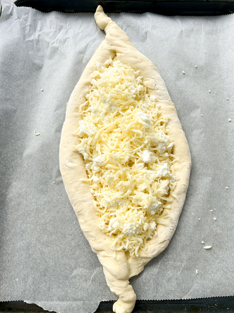 The Khachapuri's dough, in a boat shape, is placed on an oven tray lined with parchment paper and filled with three different cheeses.