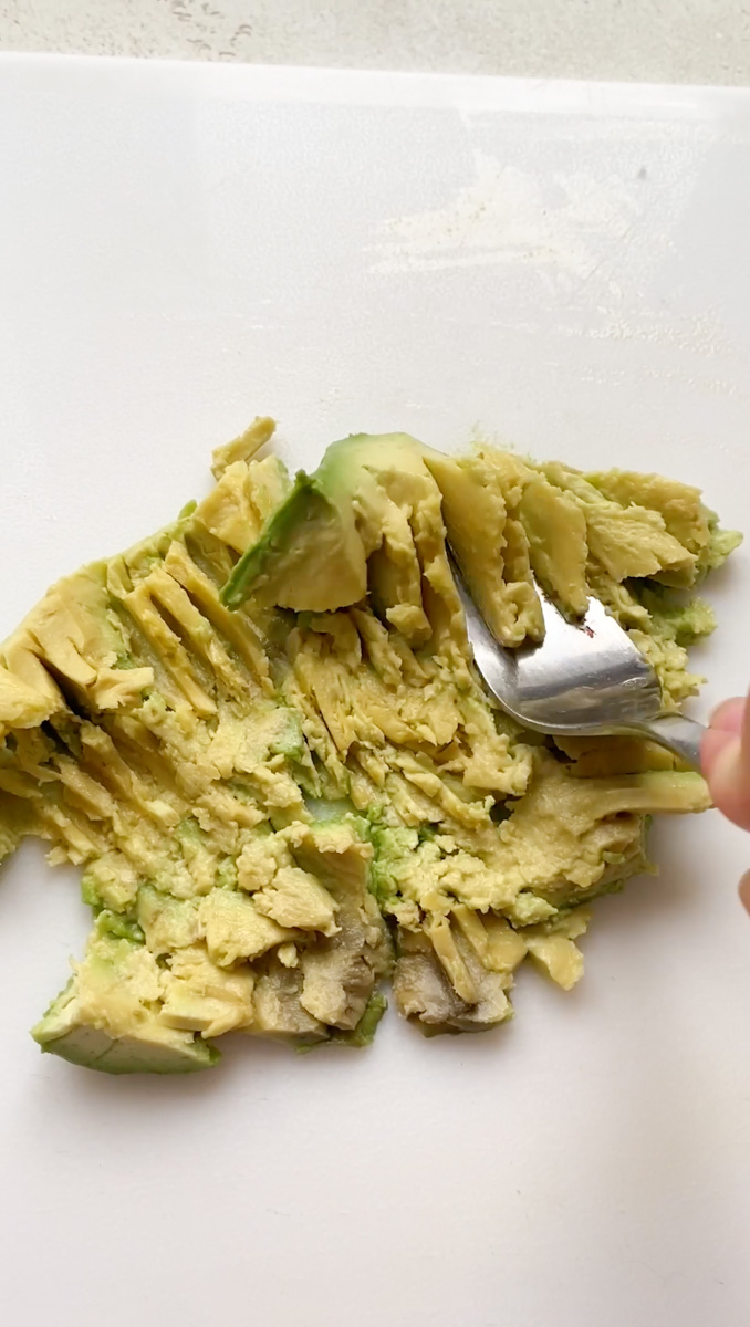 A fork mashing an avocado on a white cutting board.