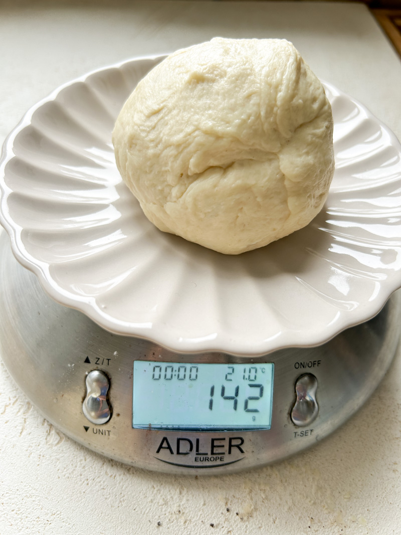 A ball of dough in a plate, on a digital kitchen scale, showing its weight is 142g.