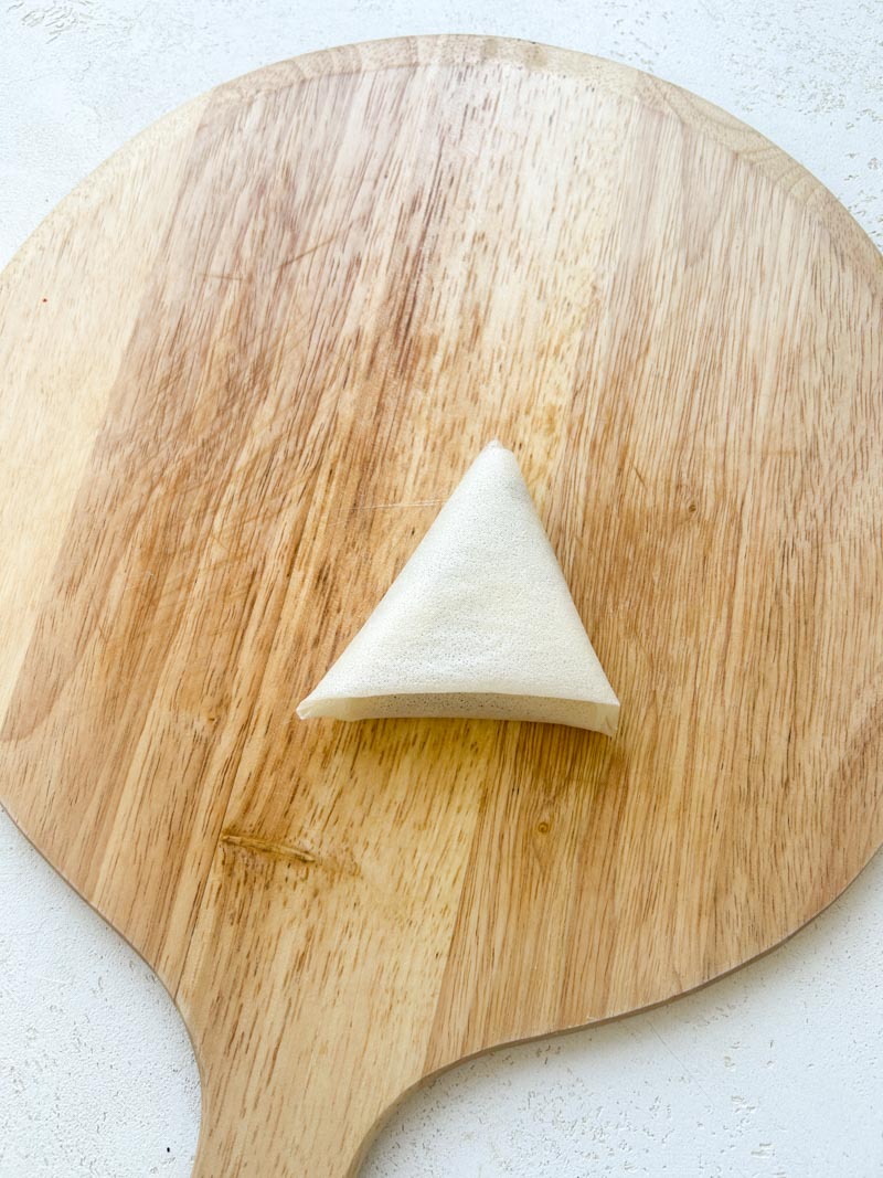 Pliage du samoussa en forme de triangle terminé. Il est posé sur la planche en bois ronde.